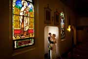 A bride and groom embrace near stained glass windows.