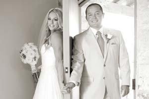 A black and white photo of a bride and groom with a door between them, holding hands.